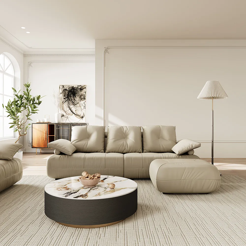 Soft beige faux leather modular sofa with deep cushions and rounded ottoman, set in a minimalist living room with a round marble coffee table and sculptural floor lamp.