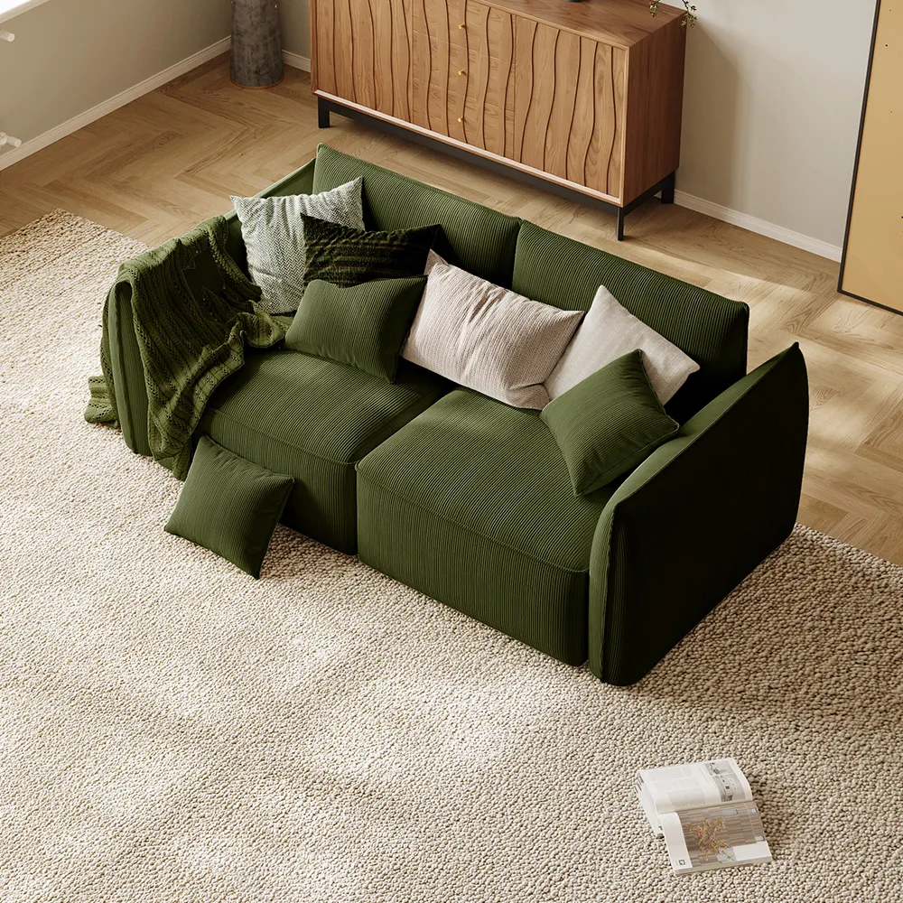 Deep green corduroy modular loveseat with layered throw pillows and a cozy knitted blanket, styled on a textured cream carpet in a warm, minimalist setting.