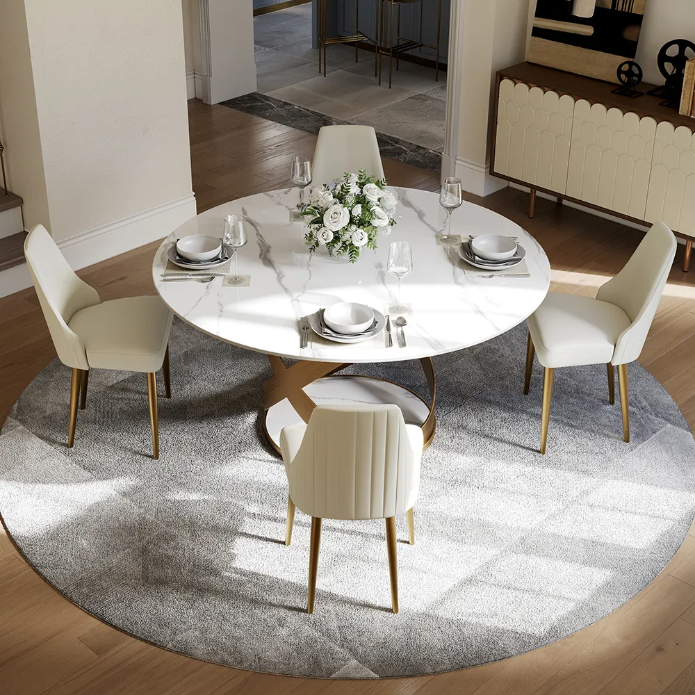 Modern round white marble dining table with gold pedestal base, paired with cream upholstered chairs and set atop a circular gray rug in a bright contemporary dining room