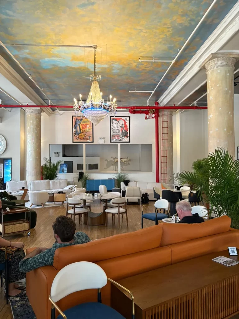 Eclectic modern showroom at NYCxDESIGN featuring contemporary leather and velvet sofas, round wood and marble coffee tables, bold pop art posters, chandelier lighting, and a hand-painted sky ceiling with classical columns—capturing the fusion of modern comfort and artistic expression.