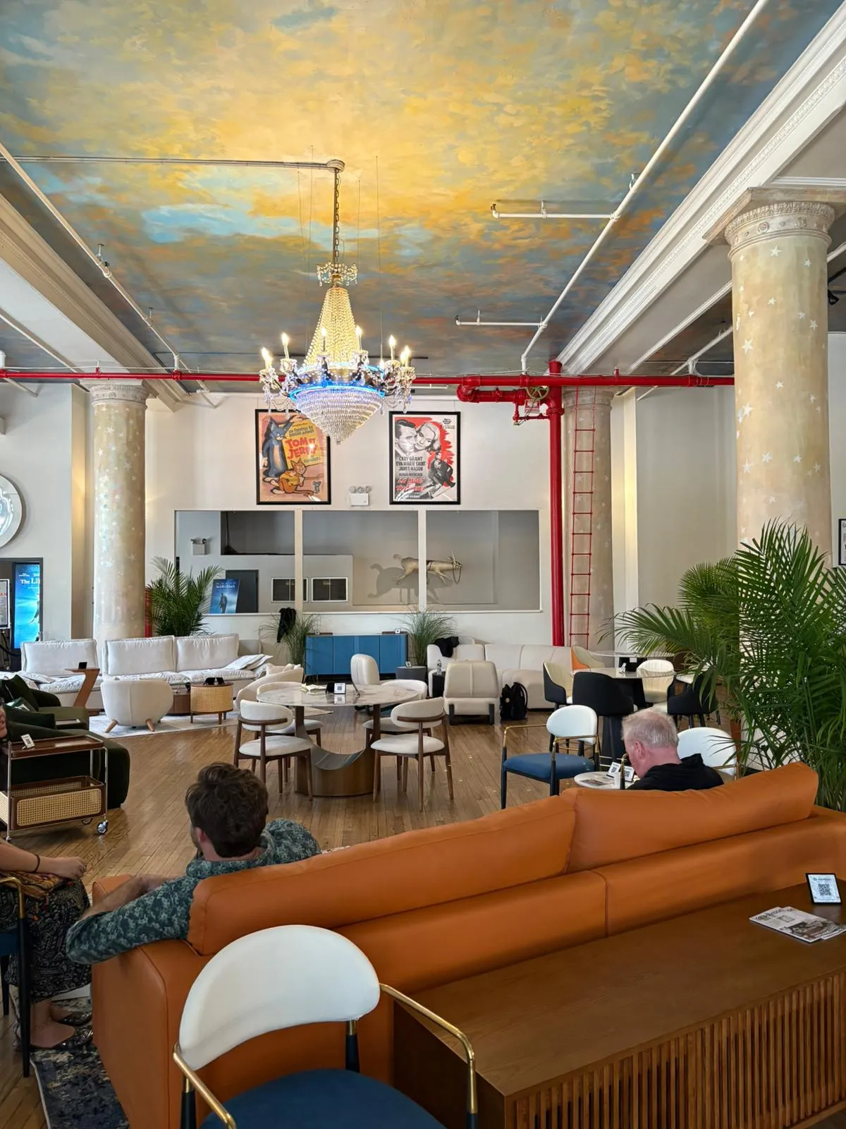 Eclectic modern showroom at NYCxDESIGN featuring contemporary leather and velvet sofas, round wood and marble coffee tables, bold pop art posters, chandelier lighting, and a hand-painted sky ceiling with classical columns—capturing the fusion of modern comfort and artistic expression.
