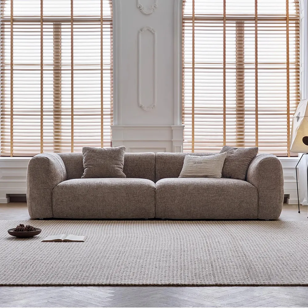 Contemporary curved-back fabric sofa in taupe color with deep seating and plush cushions, placed in a bright living room with tall windows