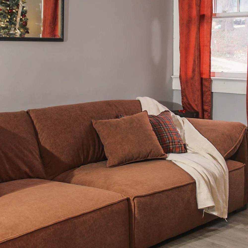 Brown L-shaped sectional sofa with plaid and beige throw pillows, placed in a cozy living room with red curtains and holiday decor.