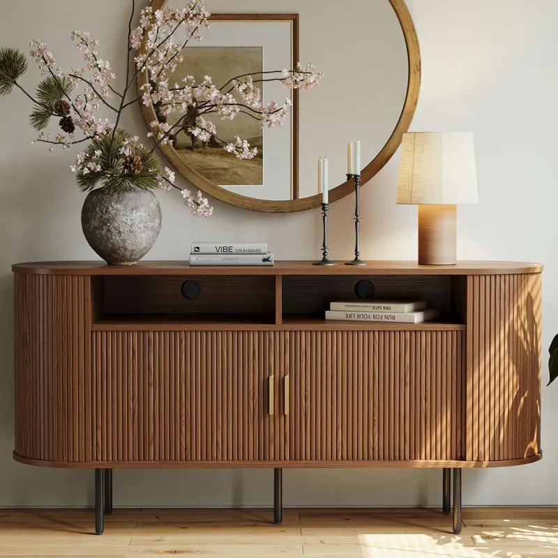 Mid-century modern fluted sideboard with open shelving and brass handles, styled with a large stone vase, table lamp, and round mirror in a neutral-toned living room.