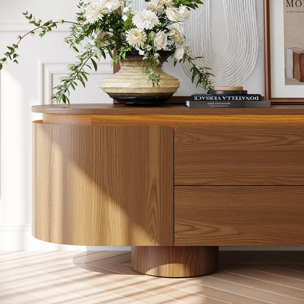 Curved walnut wood TV stand styled with floral arrangement, abstract artwork, and ceramic table lamp on top, creating a warm and elegant no-TV console display.