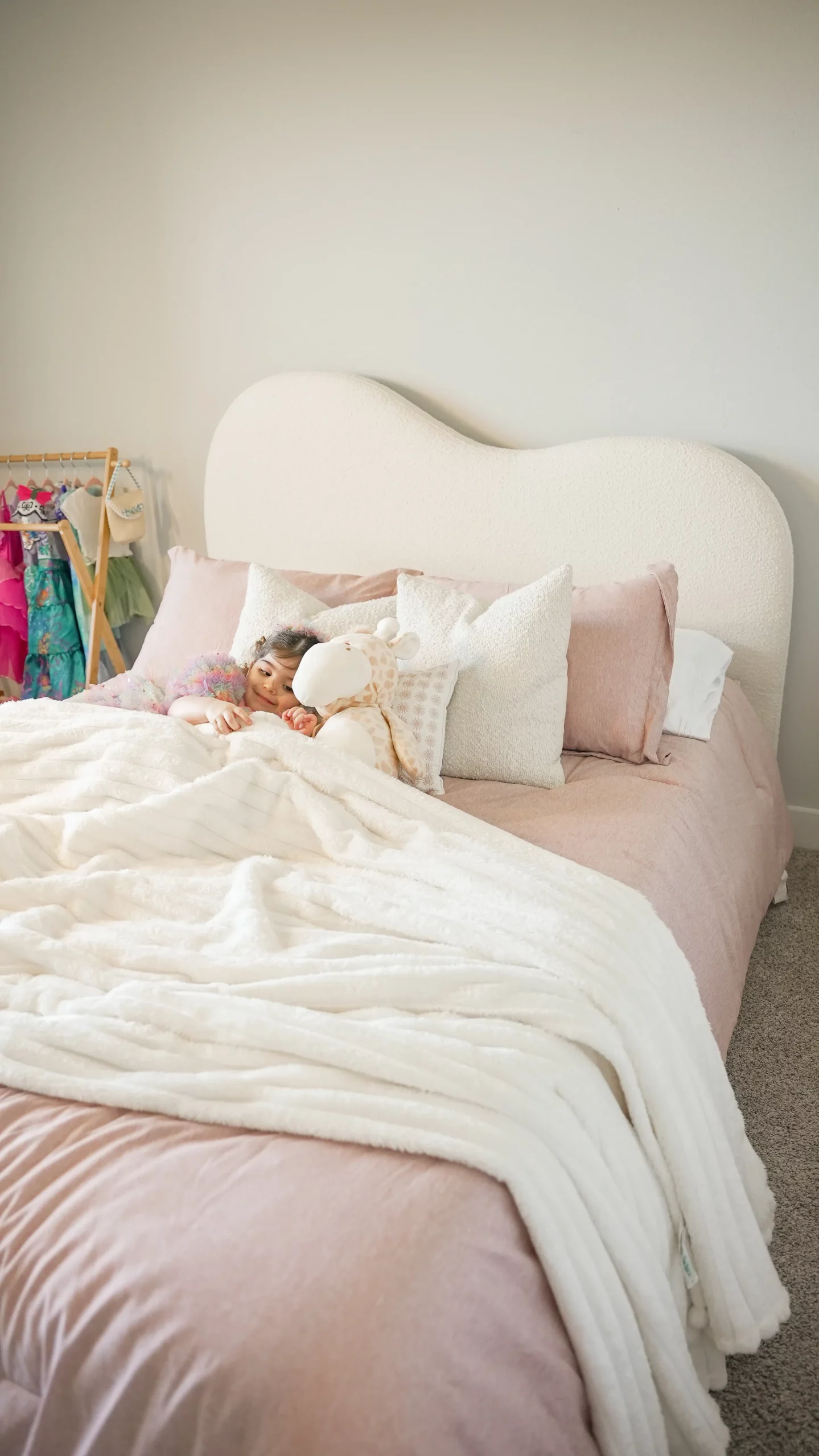 ● Young child snuggling on a boucle upholstered bed with pink bedding and plush toys in a softly lit bedroom
