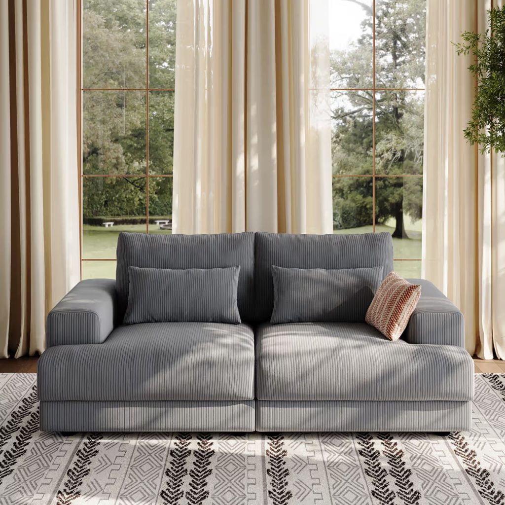 Gray corduroy modular sofa with deep cushions and wide arms in a bright living room with floor-to-ceiling windows.