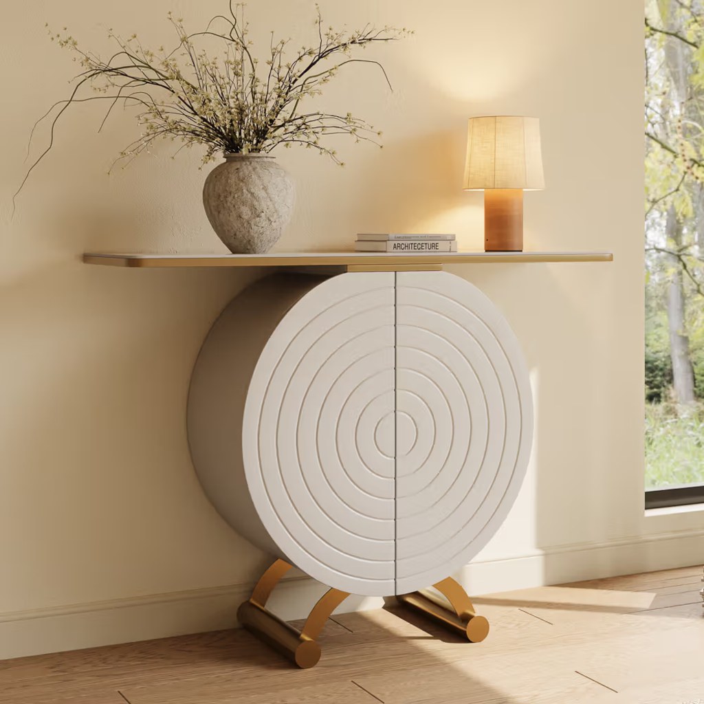 White sculptural entryway console table with circular textured doors, gold accents, a vase, books, and a table lamp.