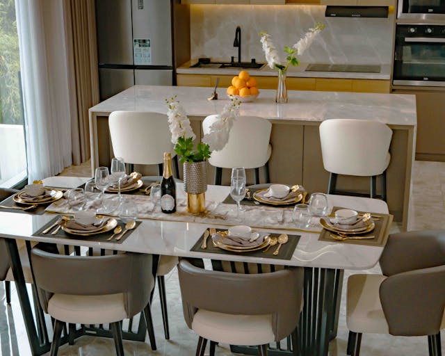 Elegant open-plan dining room with a light marble-style table, modern chairs, and a sleek kitchen backdrop.