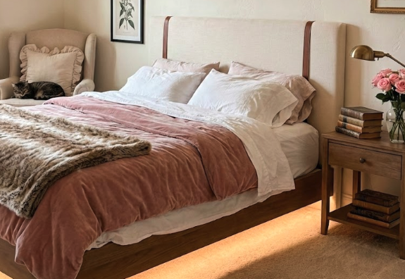Modern full-size bed with light wooden frame, layered pink velvet bedding, fur blanket, cozy armchair with cat, books, and ambient underbed lighting; visual reference for bed frame size guide.