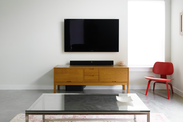 Mid-century wooden tv stand with drawers beneath a wall-mounted flat-screen TV, next to a red accent chair and glass coffee table in a bright living room.