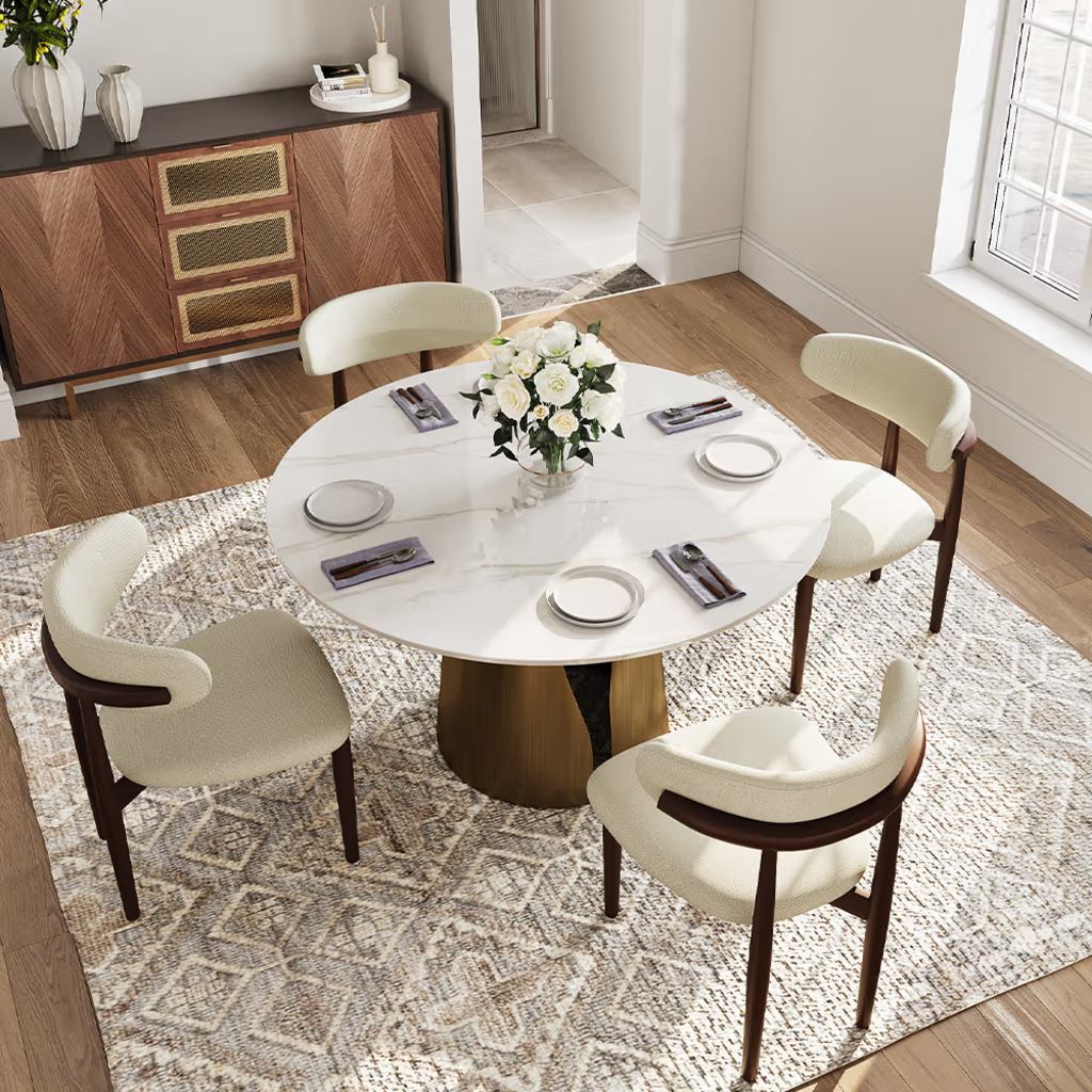 White round dining table with four cream upholstered chairs and a bronze pedestal base in a bright modern dining room.