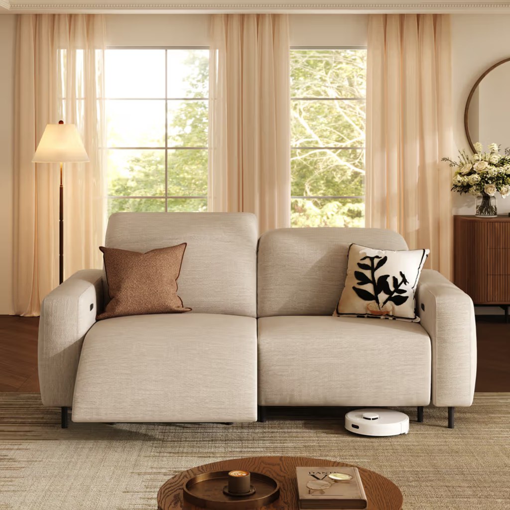 Cream power reclining loveseat with wide arms and cushioned seats in a warm living room with large windows.