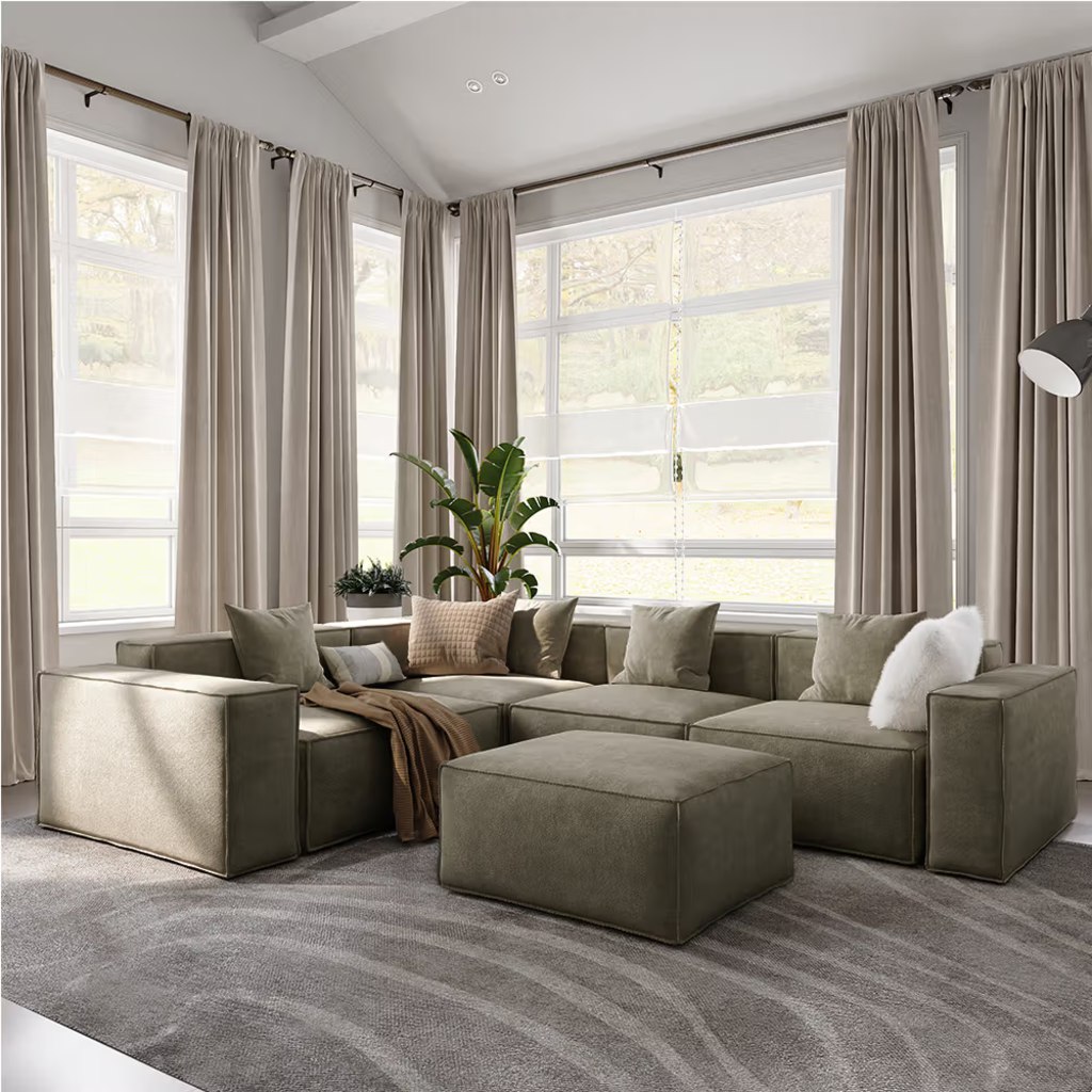 Gray modular sectional sofa with a movable ottoman in a bright living room with large corner windows.