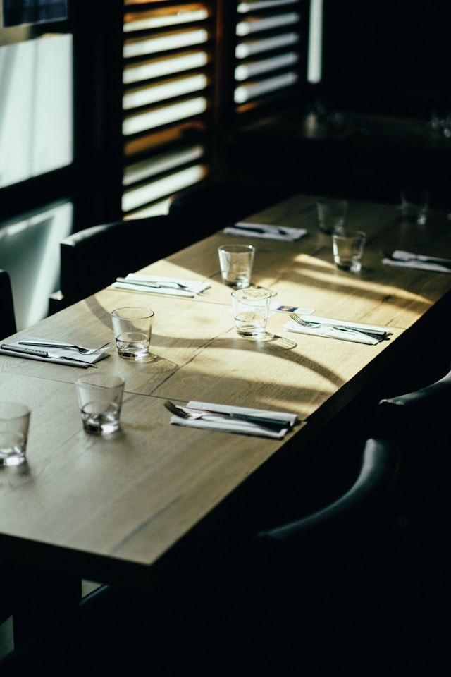 Sunlight falling across a wooden dining table set with empty glasses and cutlery in a dark, modern dining room.