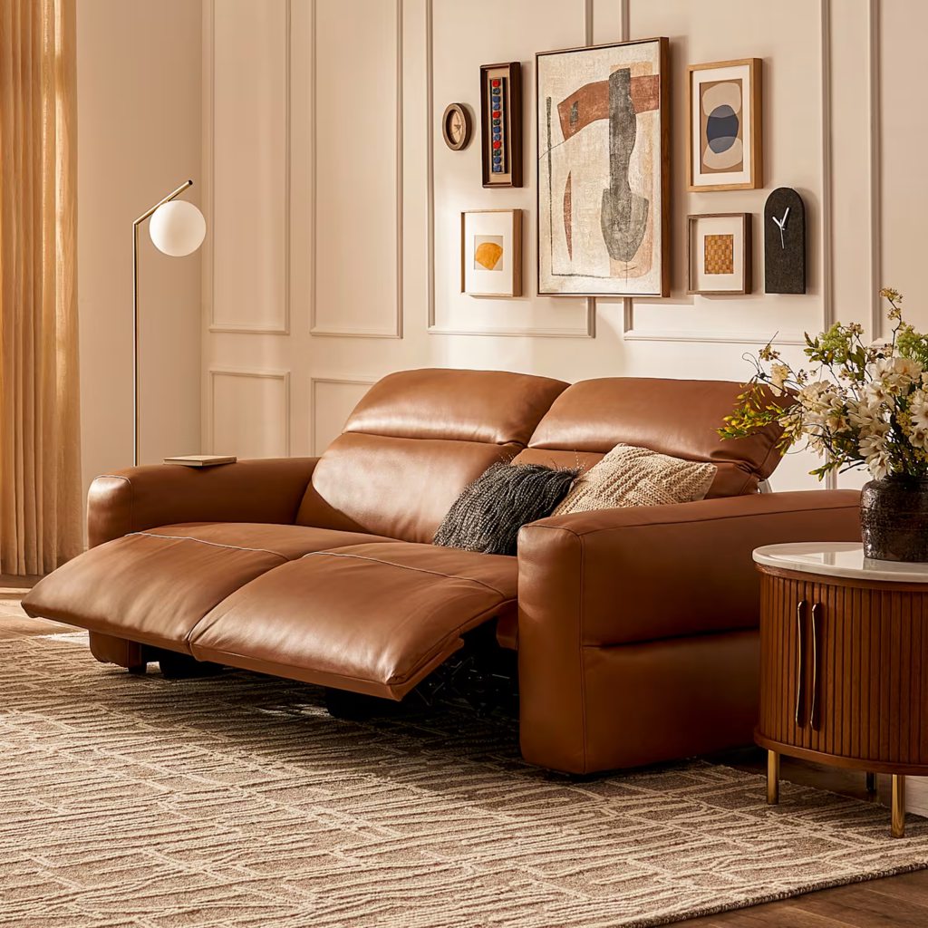 Brown leather reclining sofa with extended footrests in a warm modern living room with framed wall art, side table, and soft natural lighting.