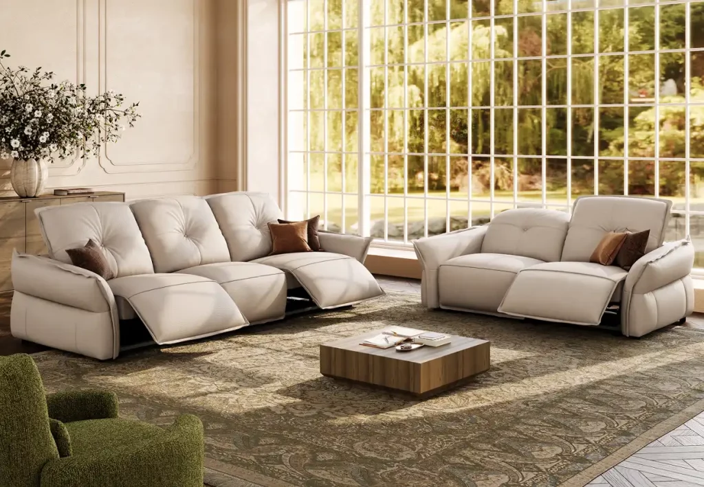 Two cream power leather reclining sofas with extended footrests and brown accent pillows in a bright living room with floor-to-ceiling windows.