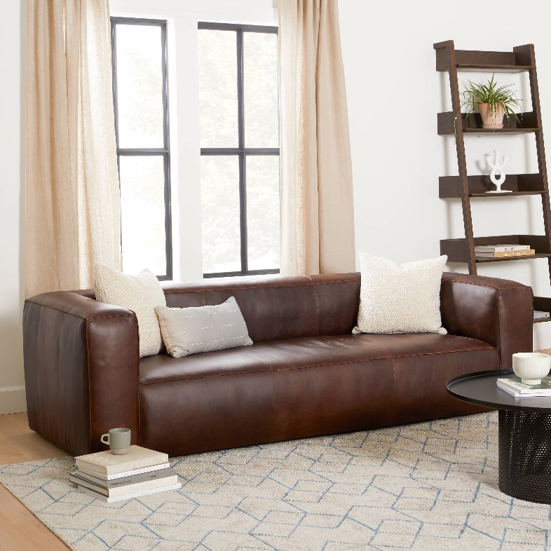 a cigar leather sofa with cushions on top near a glass coffee table in a bright and cozy living room