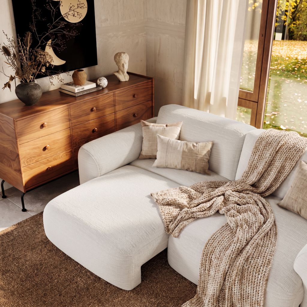 Cream sectional sofa bed with a chaise, textured throw blanket, and pillows beside a wooden dresser in a cozy, sunlit living room.
