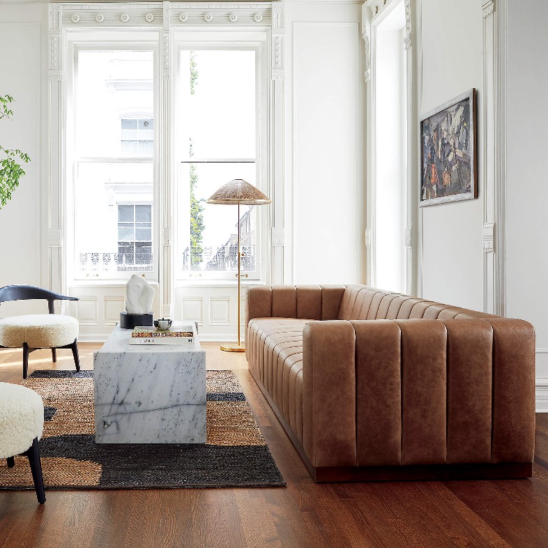 CB2 Forte 81" Channeled Saddle Leather Sofa with Walnut Base and square arms in a vintage style near a rectangular marble coffee, two wood chairs and a floor lamp in a bright and cozy living room
