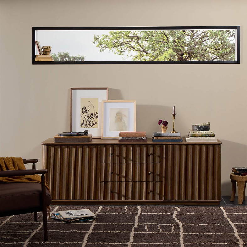 Crate & Barrel Ona 81" Fluted Wood Storage Media Console with drawers and paintings, books and a disc player on top near a chair in a bright , cozy and mid-century living room