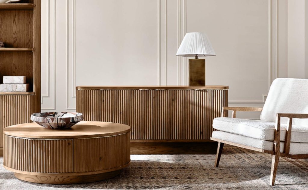 CB2 Davison 60" Fluted Oak Media Console with a lamp on top near a fluted wood shelf, a round wood fluted coffee table and a white chair in a bright and minimalist living room