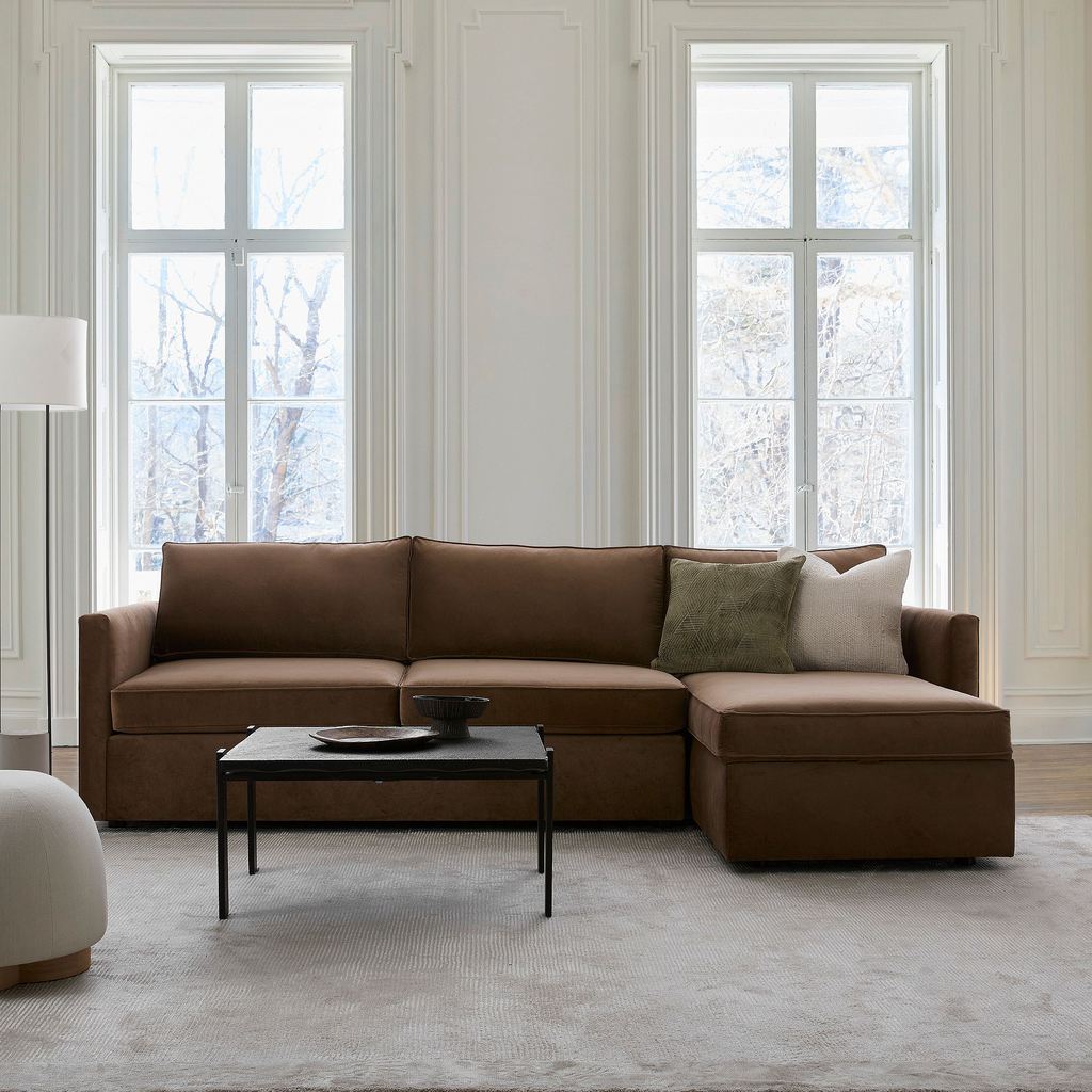 Brown chaise sleeper sectional with storage in a bright living room with tall windows and a black coffee table.