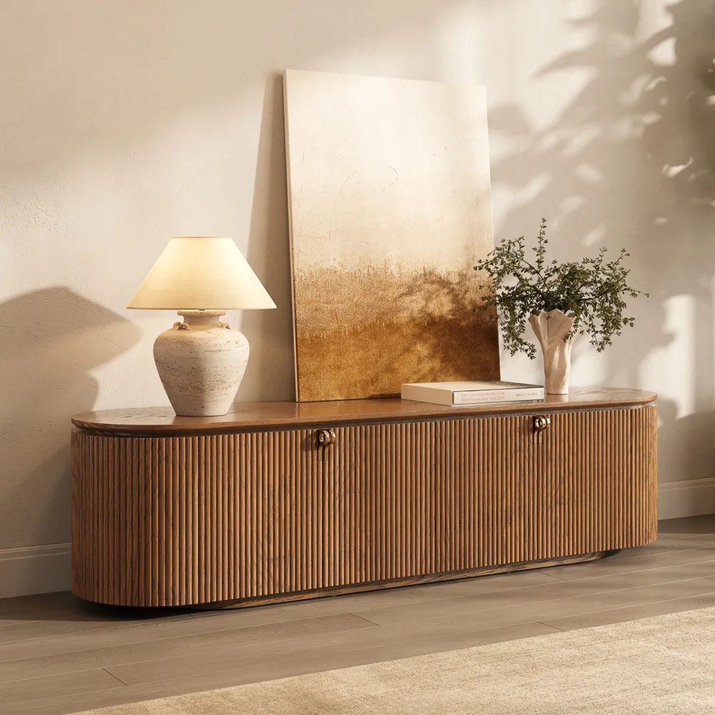 Fluted wood media console styled with a table lamp, artwork, books, and a vase of greenery.