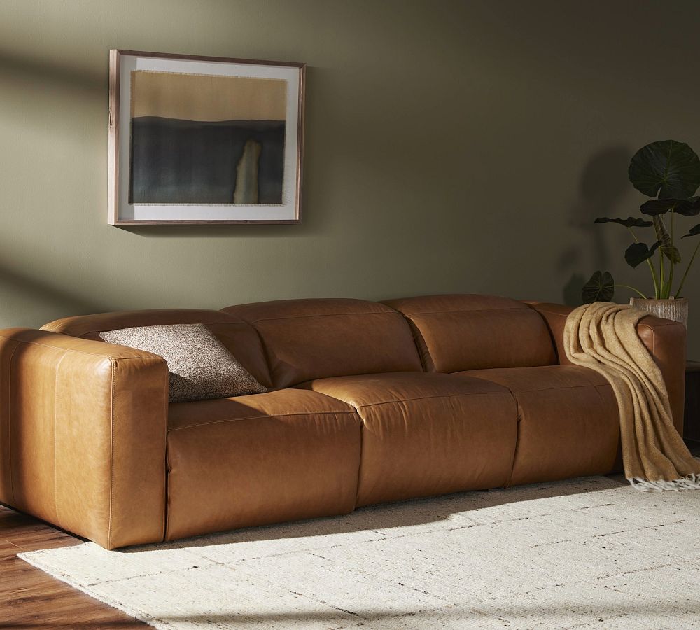 Pottery Barn Lennox 3-Piece Brown Leather Power Reclining Grand Sofa with a cushion near a plant and a mounted painting in a bright and minimalist living room