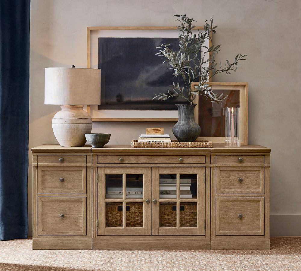 a Pottery Barn Livingston wood brown modern media console with drawers and storage for books, media and game plays and a lamp, a plant, a candle and some books on top in a bright sitting room