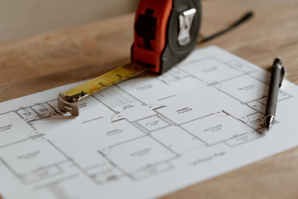 Measuring tape and pen resting on a detailed floor plan on a wooden table, ready for room measurements before buying furniture.