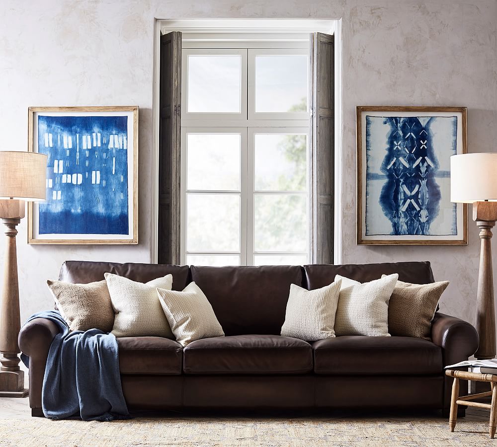 Pottery Barn Turner Roll Arm Leather Sofa in a traditional aged-leather feel with cushions near a wood chair and two floor lamps against a window in a mid-century living room