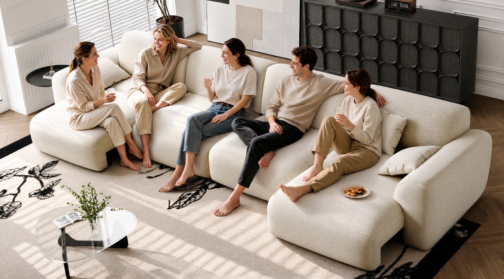 Five people relaxing and chatting on a spacious cream modular sectional sofa bed with chaise seating in a bright, modern living room.
