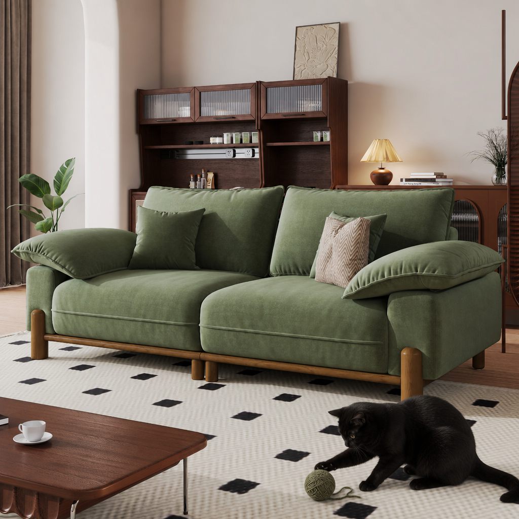 Green fabric two-seater sofa with oversized cushions and a wooden base in a cozy living room, with a black cat playing on the rug.