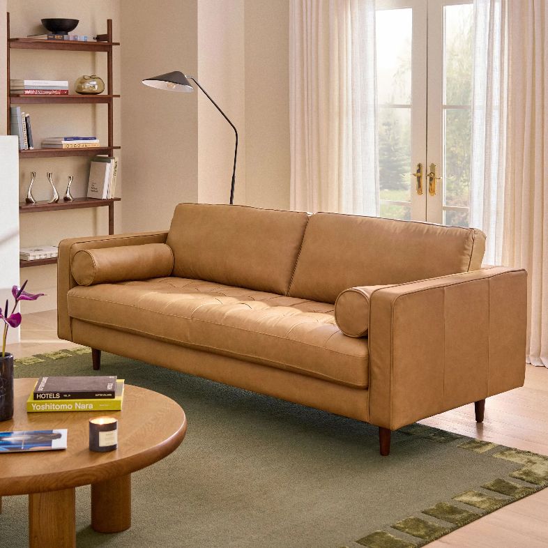 Article Sven 88" Tufted Leather Sofa with square arms and roll cushions near a round wood coffee table with books and decorations on top, a floor lamp and a bookshelf in a bright and modern living room