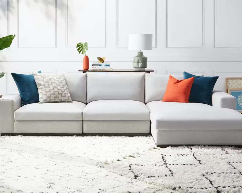 Joybird Holt Modular L Shaped Sectional with cushions in front of a wood desk with books, a plant and a lamp in a white and modern living room