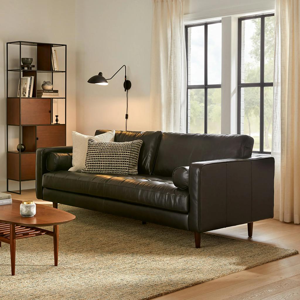 Article Sven 88" Tufted black Leather Sofa with square arms and roll cushions near an oval wood coffee table, a wood shelf and a floor lamp in front of a window in a bright and mid-century modern living room