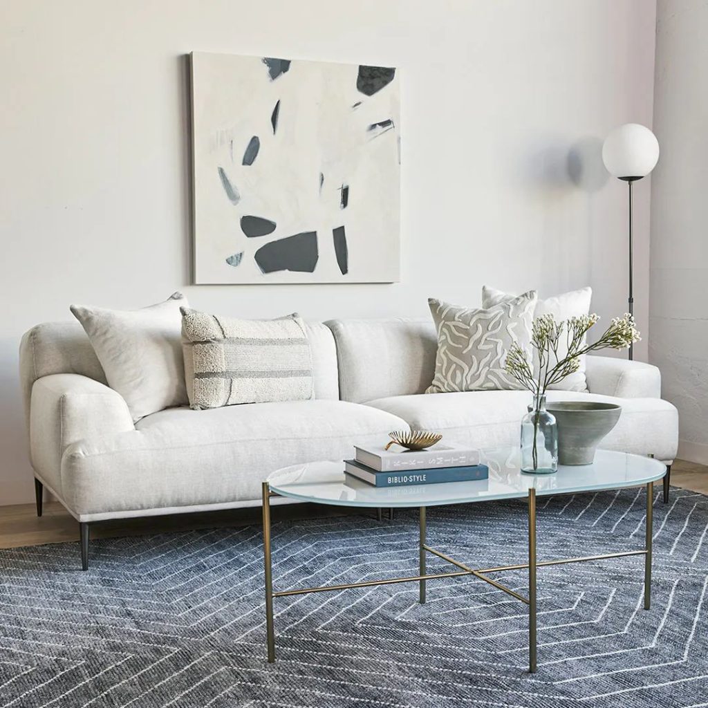 Article Abisko 91" low profile Sofa with cushions near an oval glass coffee table with books and decorations on top and a floor lamp in a minimalist and bright living room