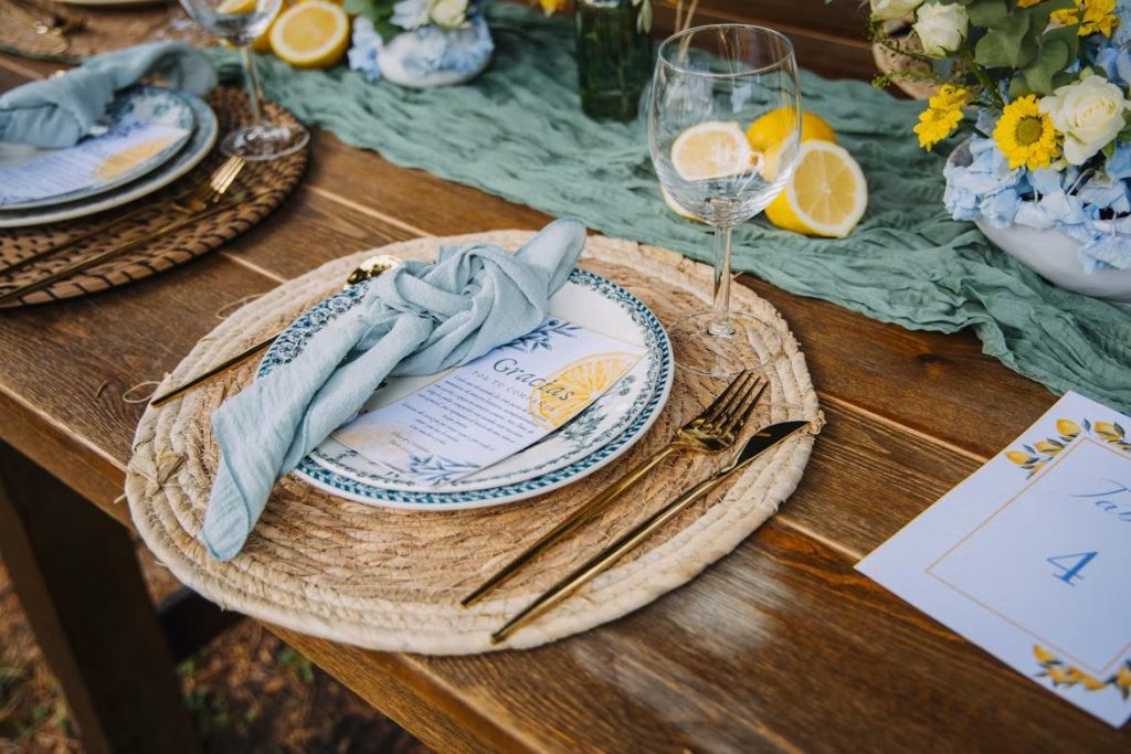 a plate, a knife, a fork and a spoon are put on a wood dining table near a glass with some decorations like flowers, lemons and table runner