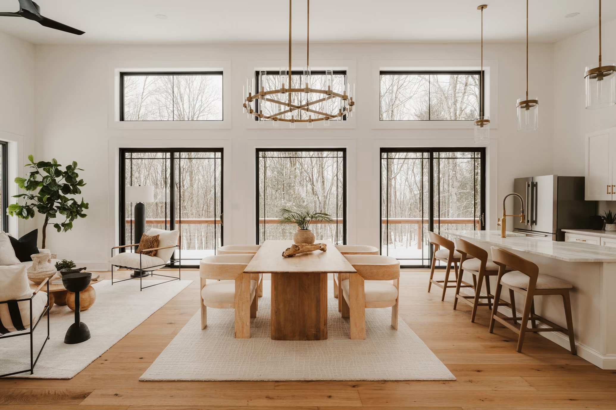 Organic modern open-plan living and dining room featuring warm wood furniture, neutral upholstery, natural light, large windows, layered textures, and a minimalist kitchen with natural materials.