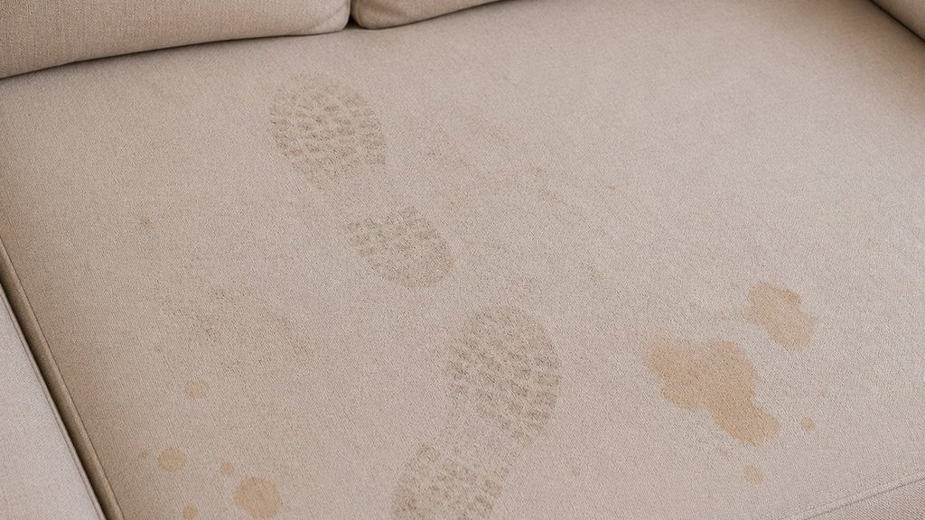Beige fabric sofa seat with visible dusty shoe prints and light stains after guests left a watch party.