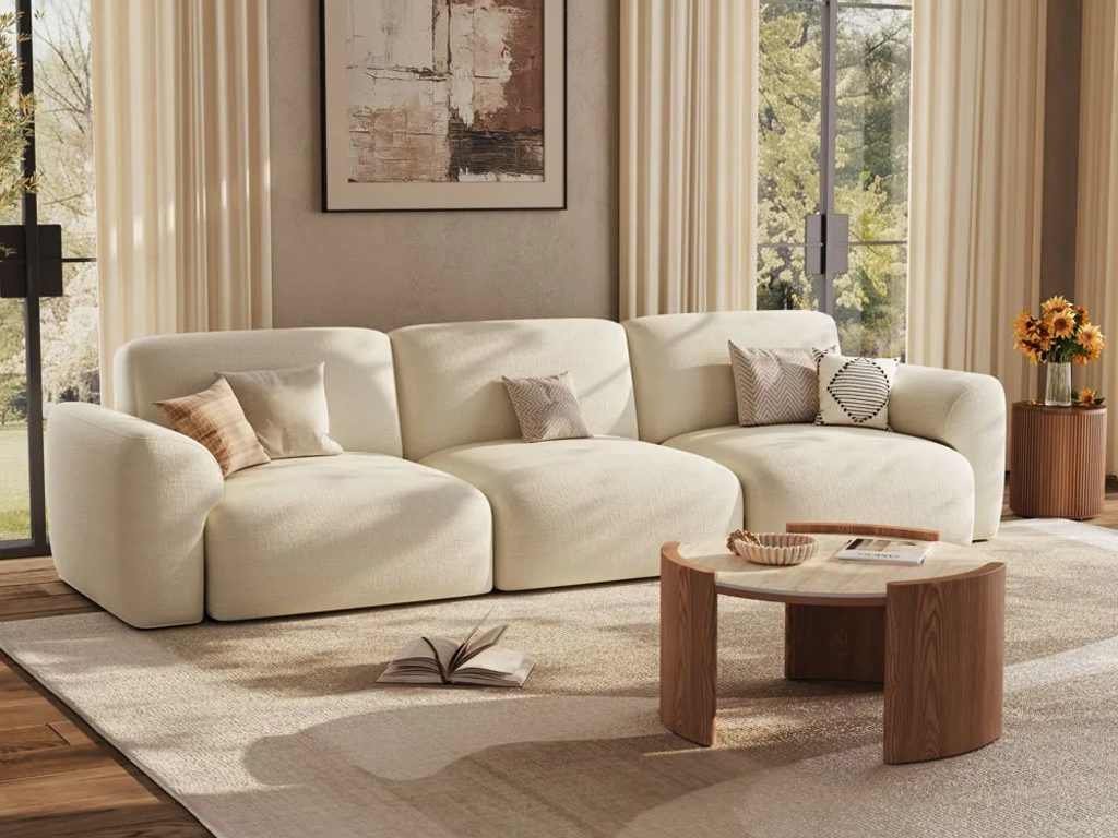 Cream curved sofa with neutral pillows, a round wood coffee table, and floor-to-ceiling windows in a warm modern living room.