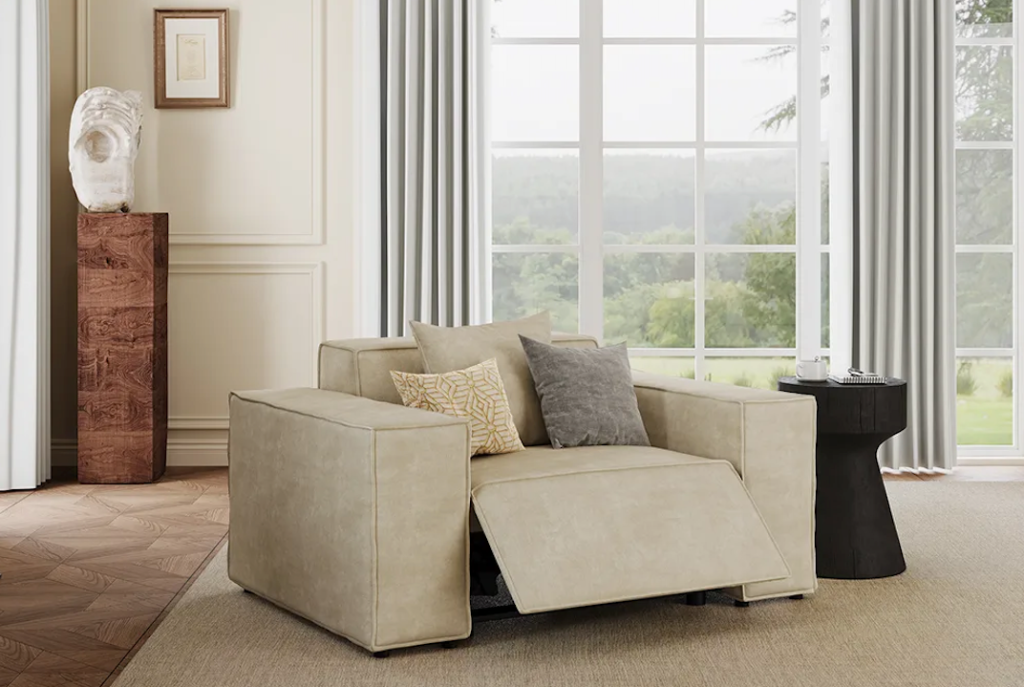A beige fabric modern power recliner armchair with its footrest partially extended by a bright window scene.