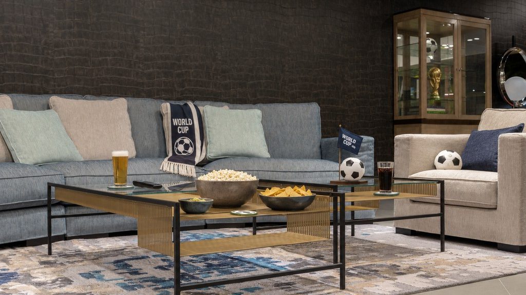 Modern living room styled for World Cup viewing with a blue sectional, soccer-themed decor, popcorn, chips, drinks, and paired glass coffee tables.