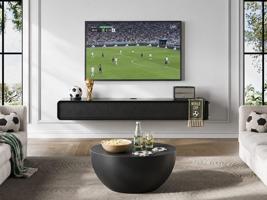Modern living room with a floating TV stand, soccer-themed pillows, World Cup decor, snacks on a round coffee table, and a live match playing on the TV.