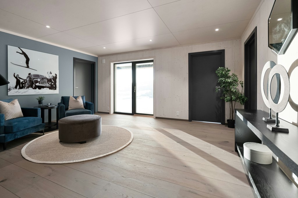 Modern sitting area with blue accent chairs, round ottoman, beige oval rug, light wood flooring, black console table, and minimalist wall decor.