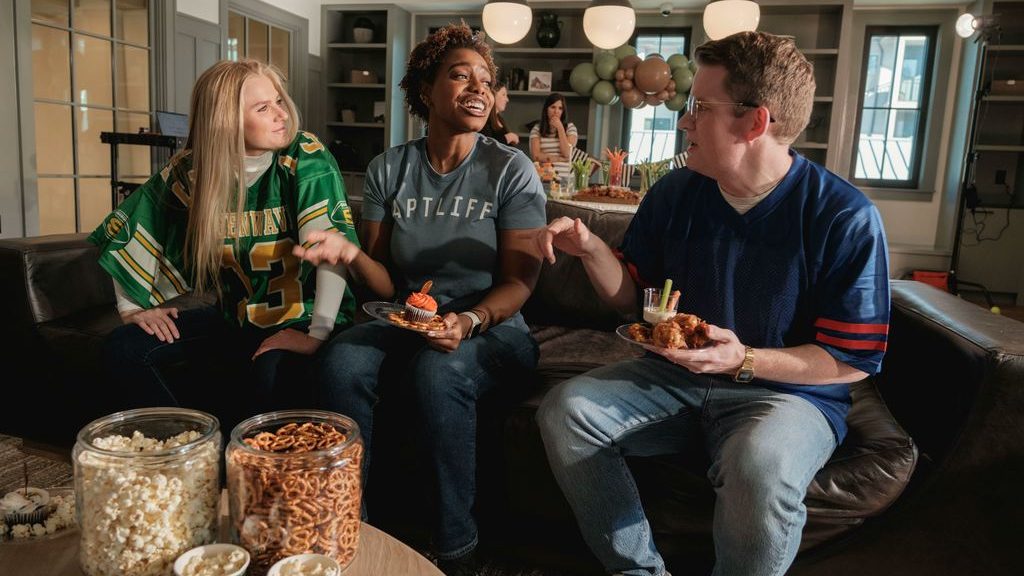 Friends in football jerseys share snacks and drinks on a sofa, with popcorn and pretzels arranged on a coffee table for a watch party.