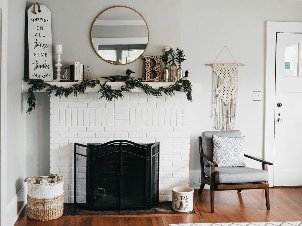 White brick fireplace with black screen, greenery garland, round mirror, woven basket, accent chair, and layered mantel decor in a cozy living room.