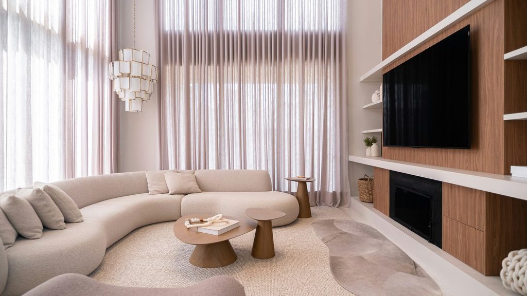 Warm modern living room with a curved cream sectional, nested wood coffee tables, floor-to-ceiling curtains, and a built-in wood TV wall.