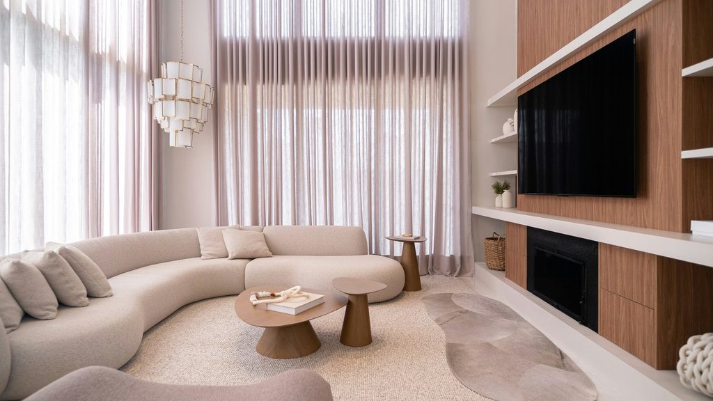 Curved cream sofa in a bright living room with tall sheer curtains, round coffee tables, wood TV wall, and soft neutral decor.